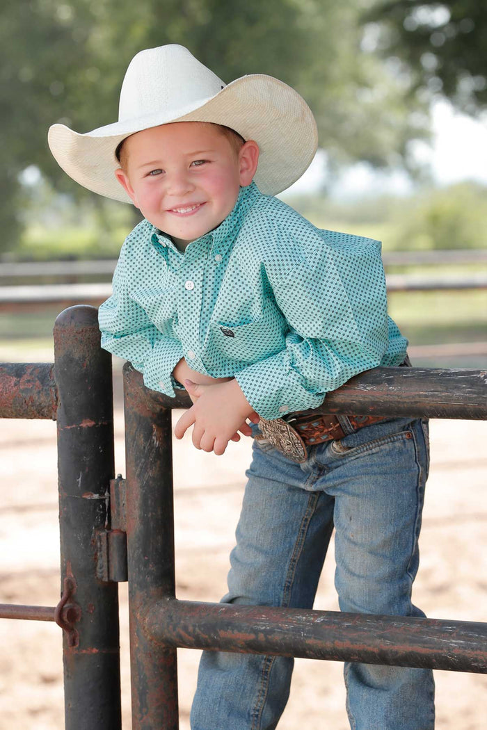 Boy leaning on a pipe fence wearing a Cinch Light Blue Geo Print Button-Down Long Sleeve Western Shirt with a straw cowboy hat, jeans, and a belt. Front view.