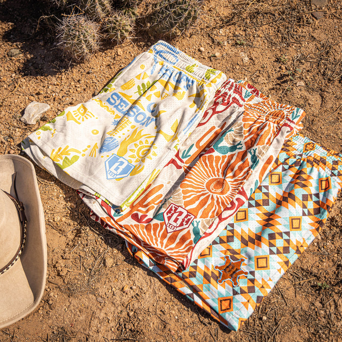 Four colorful desert patterned shorts on a sandy surface with a cowboy hat nearby