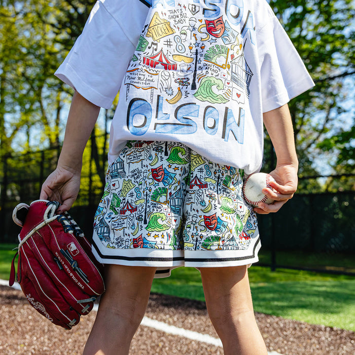 Youth player holding a ball and glove on a baseball field, showing the white Jackson Olson x BL101 Hometown Shorts paired with the Hometown Relaxed Tee back graphic