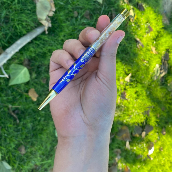 Hand holding a blue pen with gold accents against a green grass background