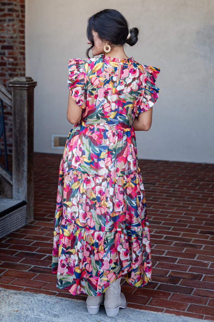 A woman viewed from behind wearing a colorful floral midi dress with ruffled short sleeves and a tiered skirt, standing on a brick patio against a stucco wall.