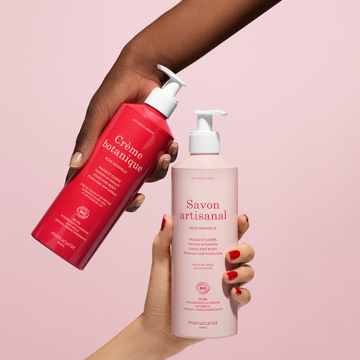 Two hands hold Manucurist’s Rose Botanical Cream Artisan Soap Duo - Cosmos Organic, featuring a red hand cream and a light pink artisan soap bottle against a pink background.