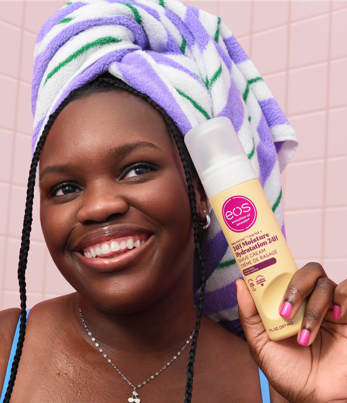 Woman with a towel on her head holding an EOS product against a pink tiled wall.