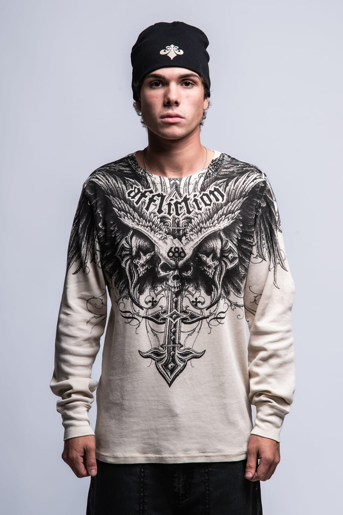 A person in a black beanie wears the 686 Affliction Death Eyes Thermal LS by 686, a long-sleeve white men's shirt with bold black graphics and "Affliction" on the chest, standing against a plain gray background.