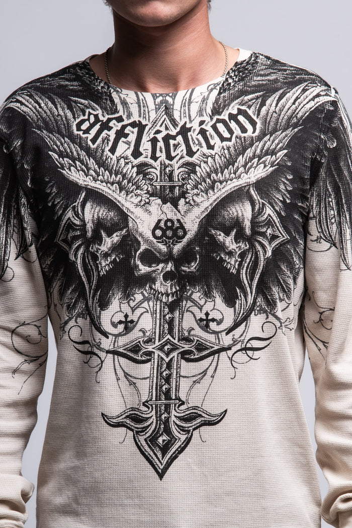 A person wears the 686 Affliction Death Eyes Thermal LS by 686‚Äîa light-colored, 100% cotton men‚Äôs tee with bold black graphics of skulls, wings, a cross, and ‚ÄúAffliction‚Äù in stylized text. Only their torso and part of neck are visible.