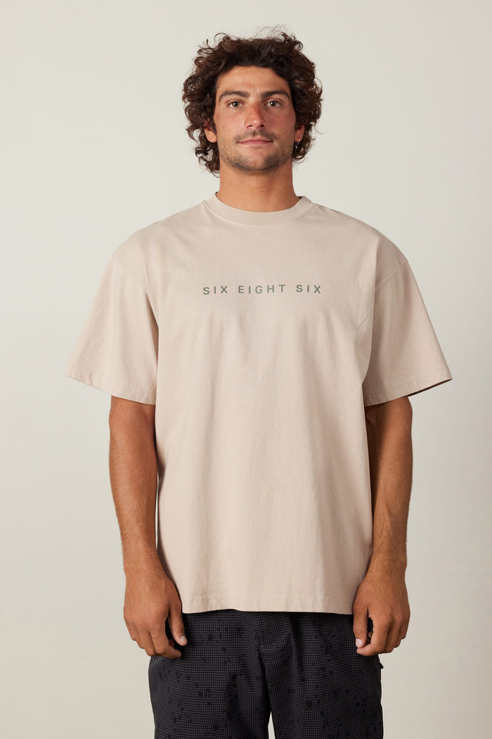 A man with curly brown hair wears the 686 Elements Relaxed S/S Tee in beige, featuring ‚ÄúSIX EIGHT SIX‚Äù on the front, paired with dark patterned shorts, standing against a plain light background.