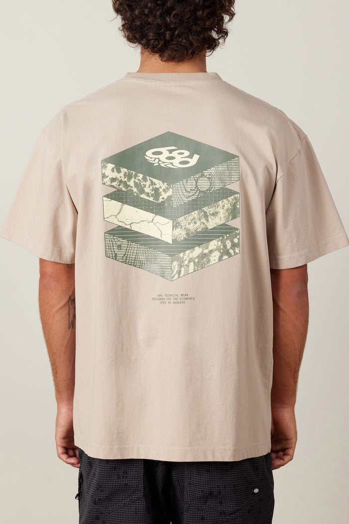A person wears the 686 Elements Relaxed S/S Tee by 686, a beige men‚Äôs t-shirt made from 100% cotton, featuring geometric hexagonal graphics and layered abstract patterns with a small text section on the back.