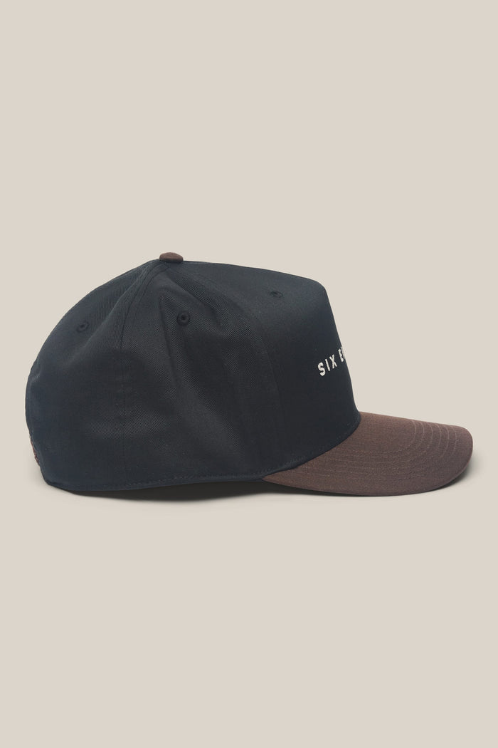 The 686 High-Pro Ses Two Tone Structured 5 Panel Hat by 686 is shown in side view. This black cotton men's hat features a brown brim, small ventilation holes, and minimal white text partially visible on the front.
