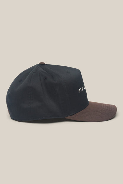 The 686 High-Pro Ses Two Tone Structured 5 Panel Hat by 686 is shown in side view. This black cotton men's hat features a brown brim, small ventilation holes, and minimal white text partially visible on the front.