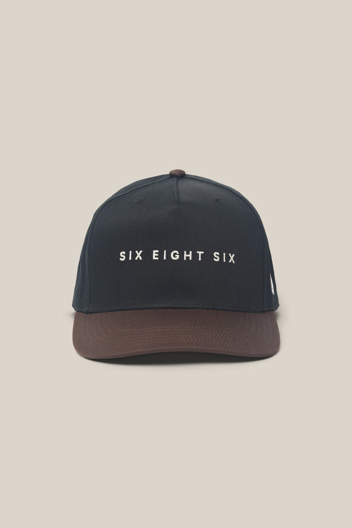 The 686 High-Pro Ses Two Tone Structured 5 Panel Hat by 686 is a black cotton hat with a brown brim, featuring "SIX EIGHT SIX" in white capital letters on the front.