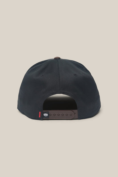 The 686 High-Pro Ses Two Tone Structured 5 Panel Hat by 686 is shown from the back, highlighting its adjustable brown strap and a small black fabric tag featuring white and red details. Made from quality cotton for lasting comfort and durability, this classic men's hat is photographed against a plain light beige background.