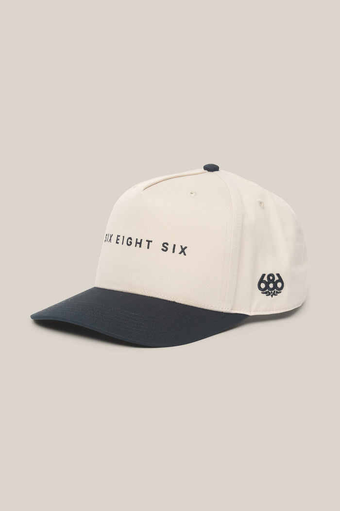 The 686 High-Pro Ses Two Tone Structured 5 Panel Hat by 686, featuring white and navy blue cotton with "SIX EIGHT SIX" printed on the front and a 686 logo on the side, is displayed on a beige background.