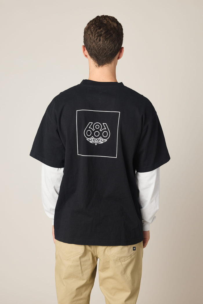 A man faces away, wearing the 686 Wordmark Relaxed 2fer LS Tee‚Äîa black cotton t-shirt with a white outlined square and 686 graphic on the back‚Äîlayered over a white long-sleeve shirt and paired with light beige pants.