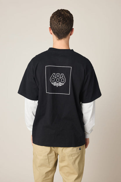 A man faces away, wearing the 686 Wordmark Relaxed 2fer LS Tee‚Äîa black cotton t-shirt with a white outlined square and 686 graphic on the back‚Äîlayered over a white long-sleeve shirt and paired with light beige pants.