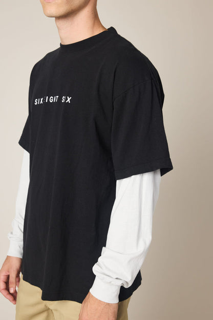 A person wearing the 686 Wordmark Relaxed 2fer LS Tee‚Äîa black men's cotton t-shirt with "SIX:EIGHT SIX" in white over long white sleeves‚Äîwith beige pants stands against a plain background, their face partially out of frame.