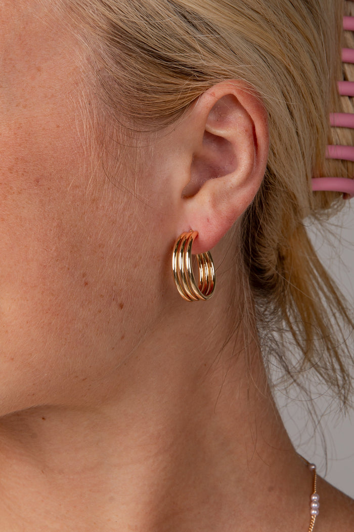 A close-up side profile of a woman with blonde hair in a pink claw clip, wearing a pair of gold triple-hoop earrings and a delicate pearl necklace against a neutral gray background.