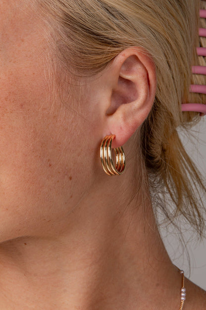 A close-up side profile of a woman with blonde hair in a pink claw clip, wearing a pair of gold triple-hoop earrings and a delicate pearl necklace against a neutral gray background.