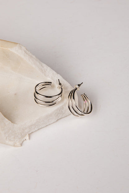 A pair of silver-toned multi-band hoop earrings resting on a textured beige stone slab against a clean white background.