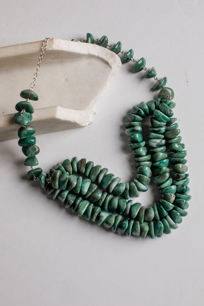 A multi-strand necklace made of polished green stone chips, draped over a light gray surface next to a white ceramic dish. The necklace features silver-toned metal chain links and a clasp.