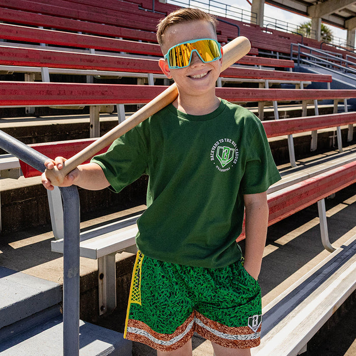 Smiling boy holding a bat on his shoulder at red stadium bleachers wearing teal shield sunglasses a forest green BL101 logo tee and green grass print mesh shorts