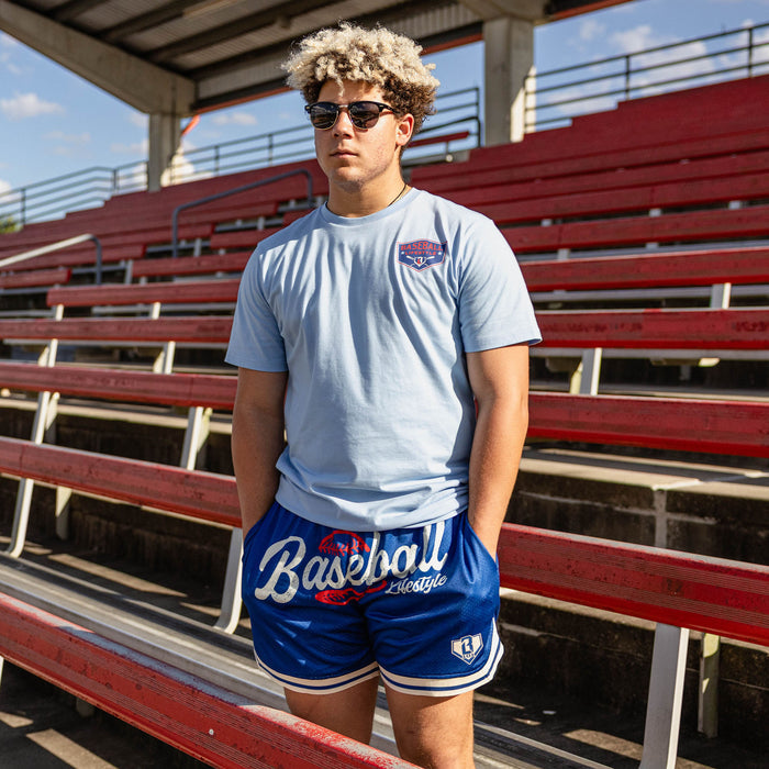 Man with curly hair in sunglasses at red stadium bleachers wearing a light blue BL101 logo tee and royal blue Baseball Lifestyle mesh shorts