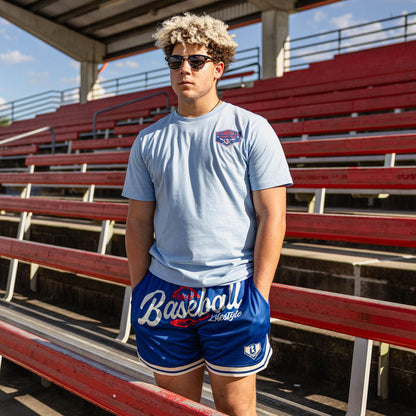 Man with curly hair in sunglasses at red stadium bleachers wearing a light blue BL101 logo tee and royal blue Baseball Lifestyle mesh shorts