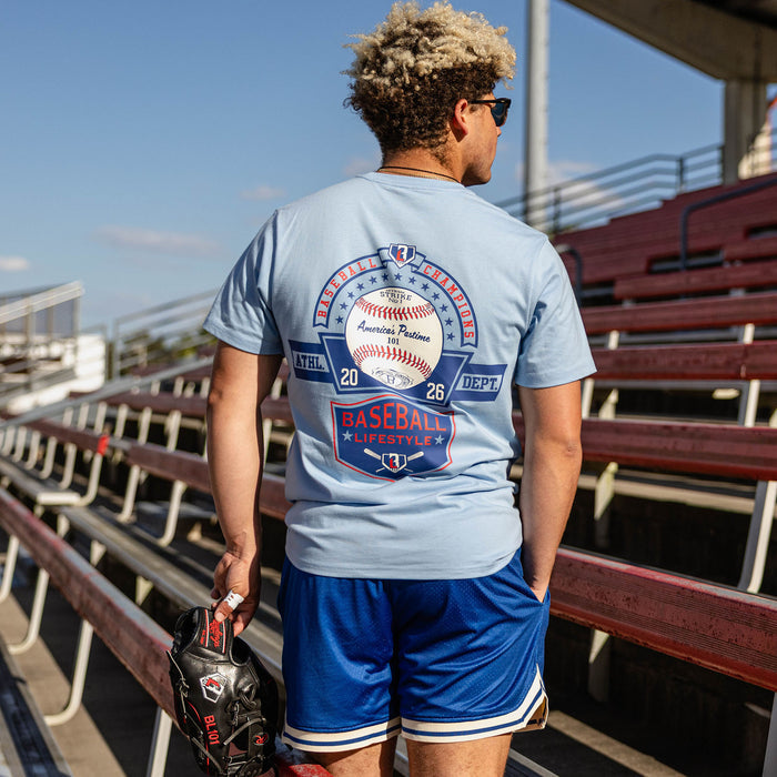 Back view of man holding a glove at red stadium bleachers wearing a light blue tee with Baseball Champions America's Pastime 101 back text and royal blue mesh shorts