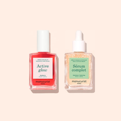 Two bottles from Manucurist’s Basic Routine - Sensitive Nails—a vibrant red Active Glow nail polish with a white cap and a clear Serum Complet cuticle serum with a dropper—are beautifully arranged on a light peach background, offering the perfect duo for nail care and moisturized nails.