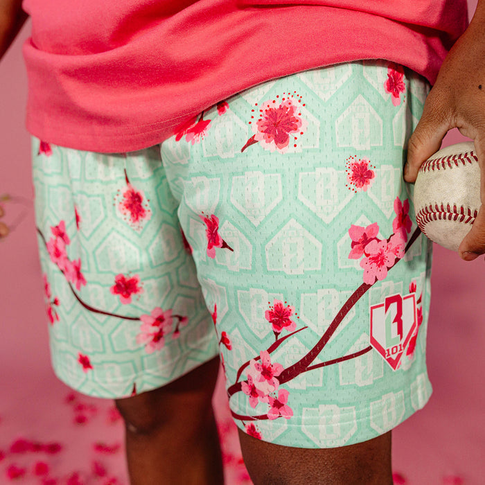 Close up of pink cherry blossom print and BL101 shield logo on mint green mesh shorts with home plate background pattern, worn by a male model holding a baseball