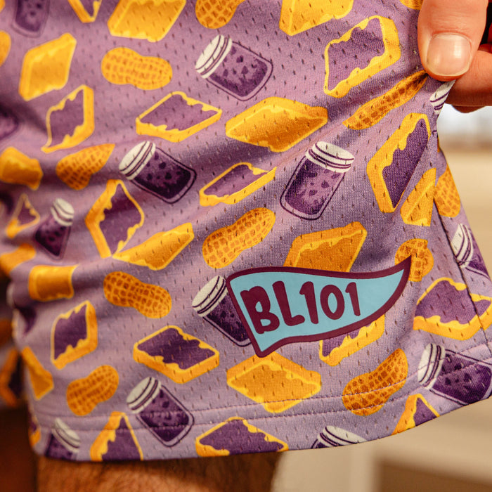 Detail close-up of the BL101 pennant logo on the purple PB&J print mesh shorts, highlighting the all-over sandwich and peanut graphics.