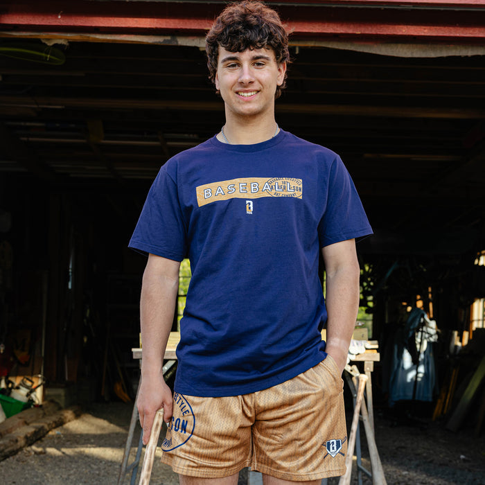 Young adult male model smiling outside a barn wearing the brown BL101 Bat Co Shorts paired with the navy Radar Tee and holding a wooden baseball bat.