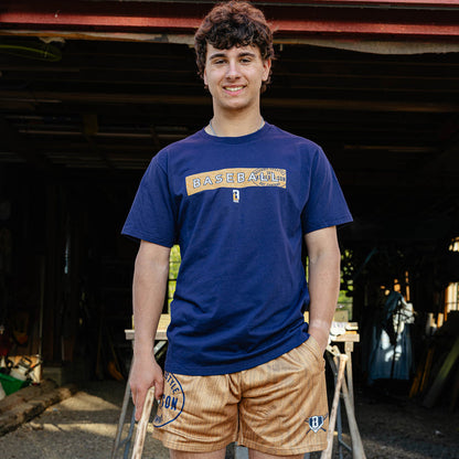 Young adult male model smiling outside a barn wearing the brown BL101 Bat Co Shorts paired with the navy Radar Tee and holding a wooden baseball bat.