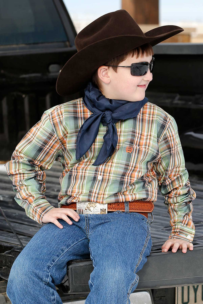 Little boy wearing green/orange/black plaid Cinch long sleeve shirt. He is also wearing sunglasses, a black cowboy hat, jeans, and a belt. Front view of him seated on the tailgate of a pickup.