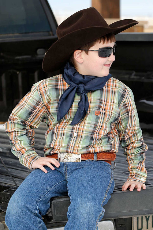 Little boy wearing green/orange/black plaid Cinch long sleeve shirt. He is also wearing sunglasses, a black cowboy hat, jeans, and a belt. Front view of him seated on the tailgate of a pickup.