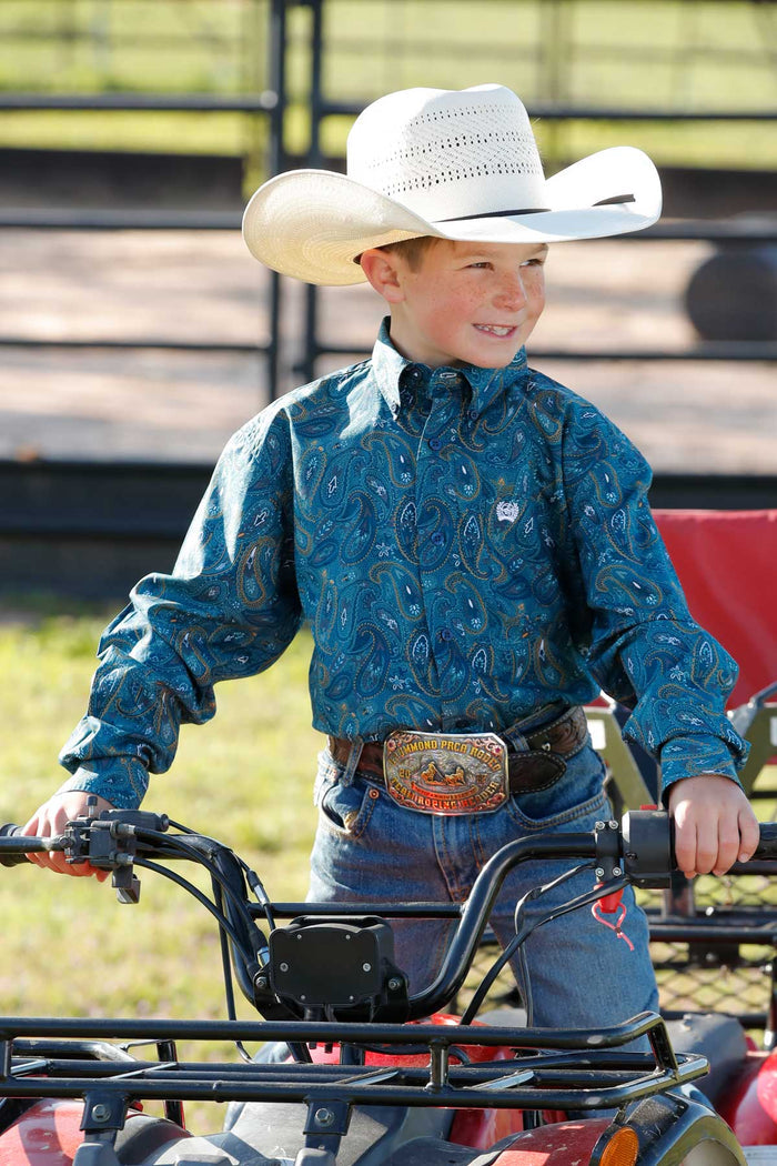 Little boy sitting on a 4-wheeler wearing a Cinch Teal Blue Paisley Print Button Down Western Shirt with a straw hat, jeans, and a belt buckle. Front view.