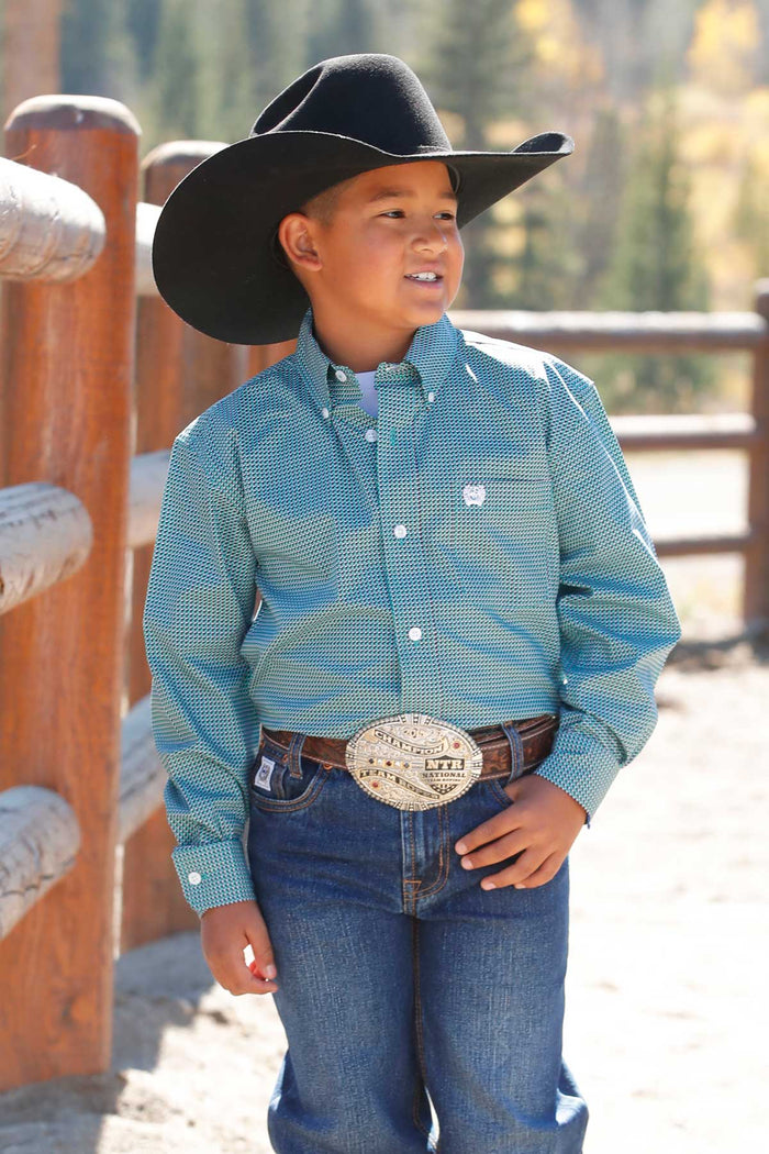 Little boy wearing turquoise with geo print Cinch long sleeve shirt. He is also wearing a black cowboy hat, jeans, and a belt. Front view of him standing beside a wooden pole fence.