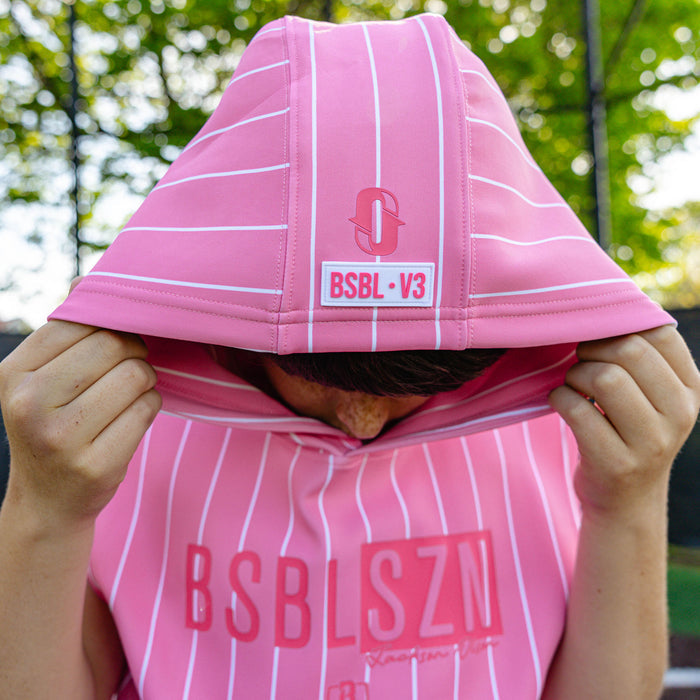 Youth player pulling up the hood of the pink pinstripe BSBL-SZN Short Sleeve Hoodie V3, showing the Jackson Olson "O" logo and "BSBL·V3" label