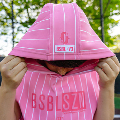 Youth player pulling up the hood of the pink pinstripe BSBL-SZN Short Sleeve Hoodie V3, showing the Jackson Olson "O" logo and "BSBL·V3" label