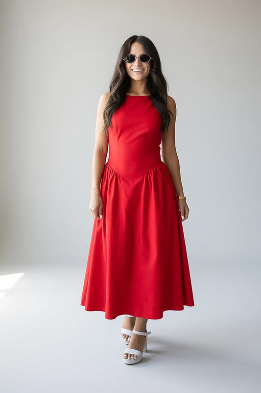 A woman modeling a sleeveless red midi dress with a fitted bodice and flared skirt, paired with white heeled sandals and sunglasses, standing against a plain light gray studio background.