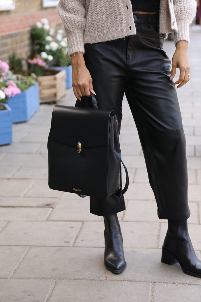 Female model holding Casey Black faux leather Laptop Backpack below her waist by the top reptile printed grab handle
