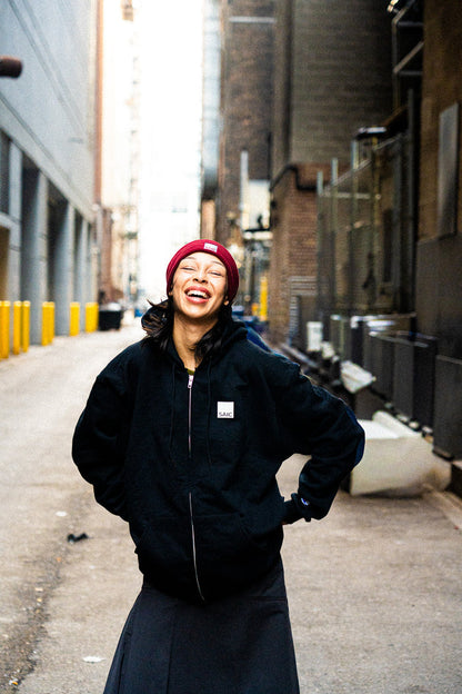 Person wearing a black jacket and red beanie standing in an urban alleyway.