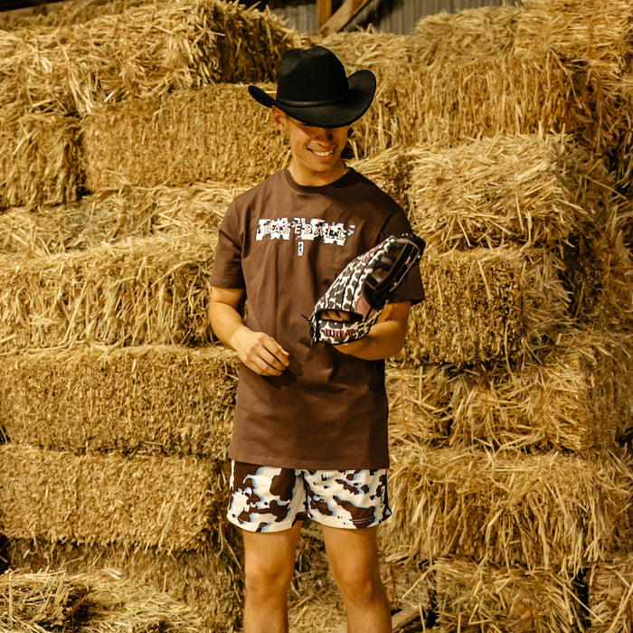 Person wearing a cowboy hat,  cow themed shorts and brown t-shirt standing in front of hay bales
