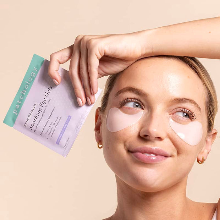 Smiling blonde woman wearing translucent pink gel patches under her eyes, arm resting on her head as she holds up a lavender Soothing Eye Gels sachet against a peach backdrop.