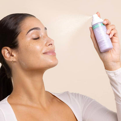 Freckled woman with dark hair pulled back, eyes closed and smiling, spritzing a fine cloud of Calm & Protect Micro Mist toward her dewy face against a warm beige backdrop.