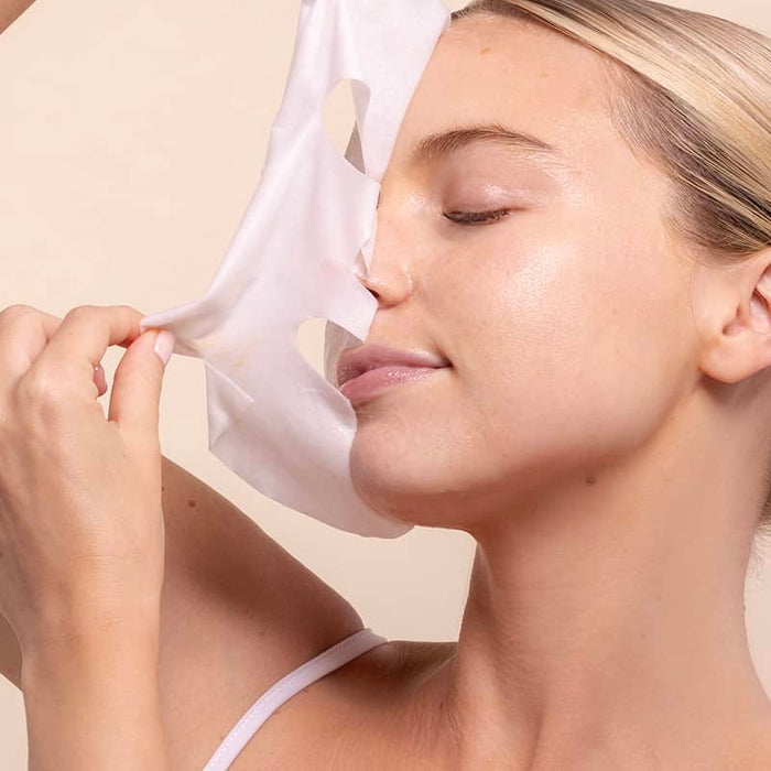 Close-up of a blonde woman with eyes closed peeling a white sheet mask away from her face to reveal dewy, glowing skin underneath, set against a soft peach backdrop.