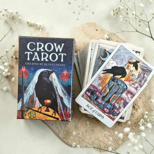 Crow Tarot card set with deck and box on a light surface with flowers.