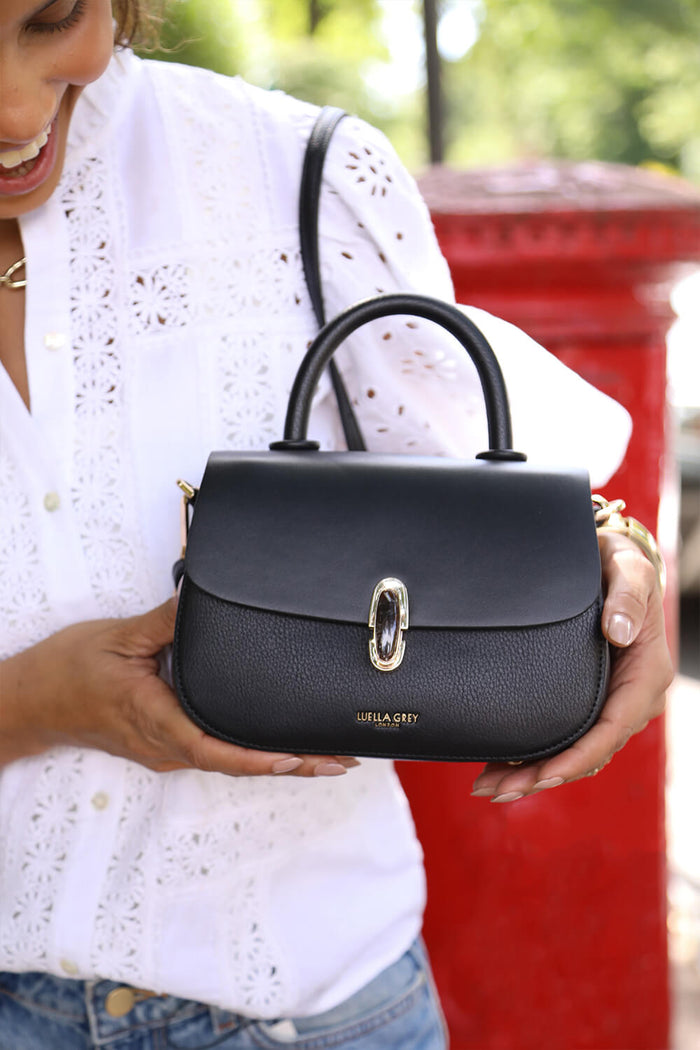 Female model holding the Delia Black faux leather Top Handle Crossbody by the bottom of the bag showing the front