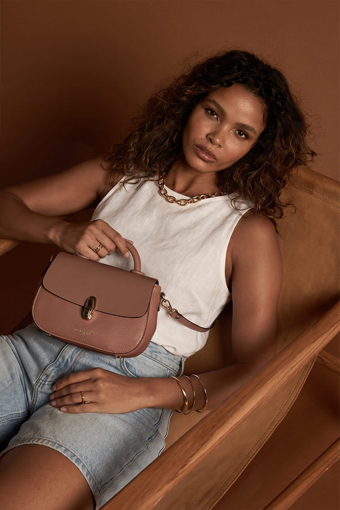 Female model sitting down holding the Delia Pecan faux leather Top Handle Crossbody by the bag's top grab handle showing the front