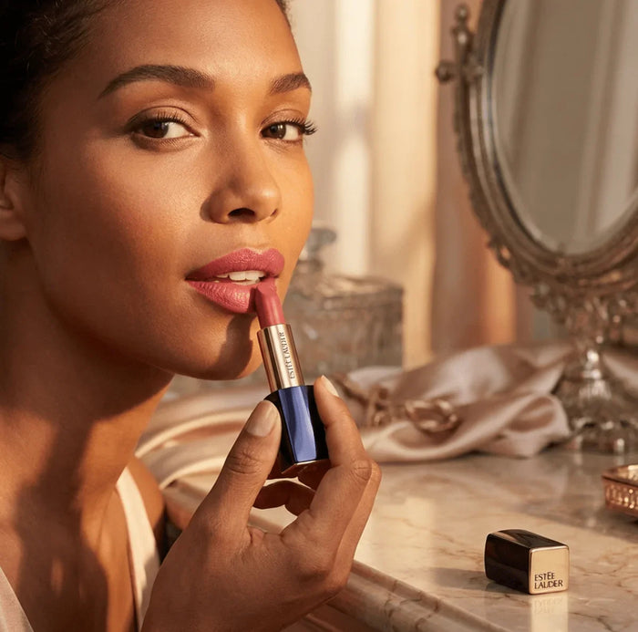 Woman applying pink matte lipstick in elegant vanity setting with gold mirror