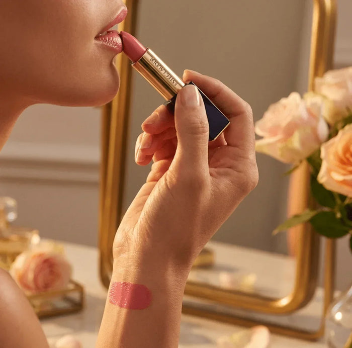 Woman applying pink matte lipstick, with lipstick swatch on wrist, near roses and a mirror.